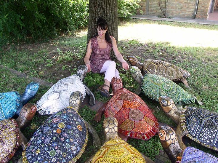 A woman sits on grass under a tree, surrounded by large, colorful, mosaic-tiled tortoise sculptures.