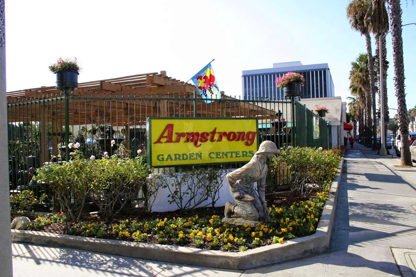 Entrance to Armstrong Garden Centers with a statue of a kneeling gardener, flower beds, and a pergola, set along a city sidewalk with palm trees and nearby office buildings.