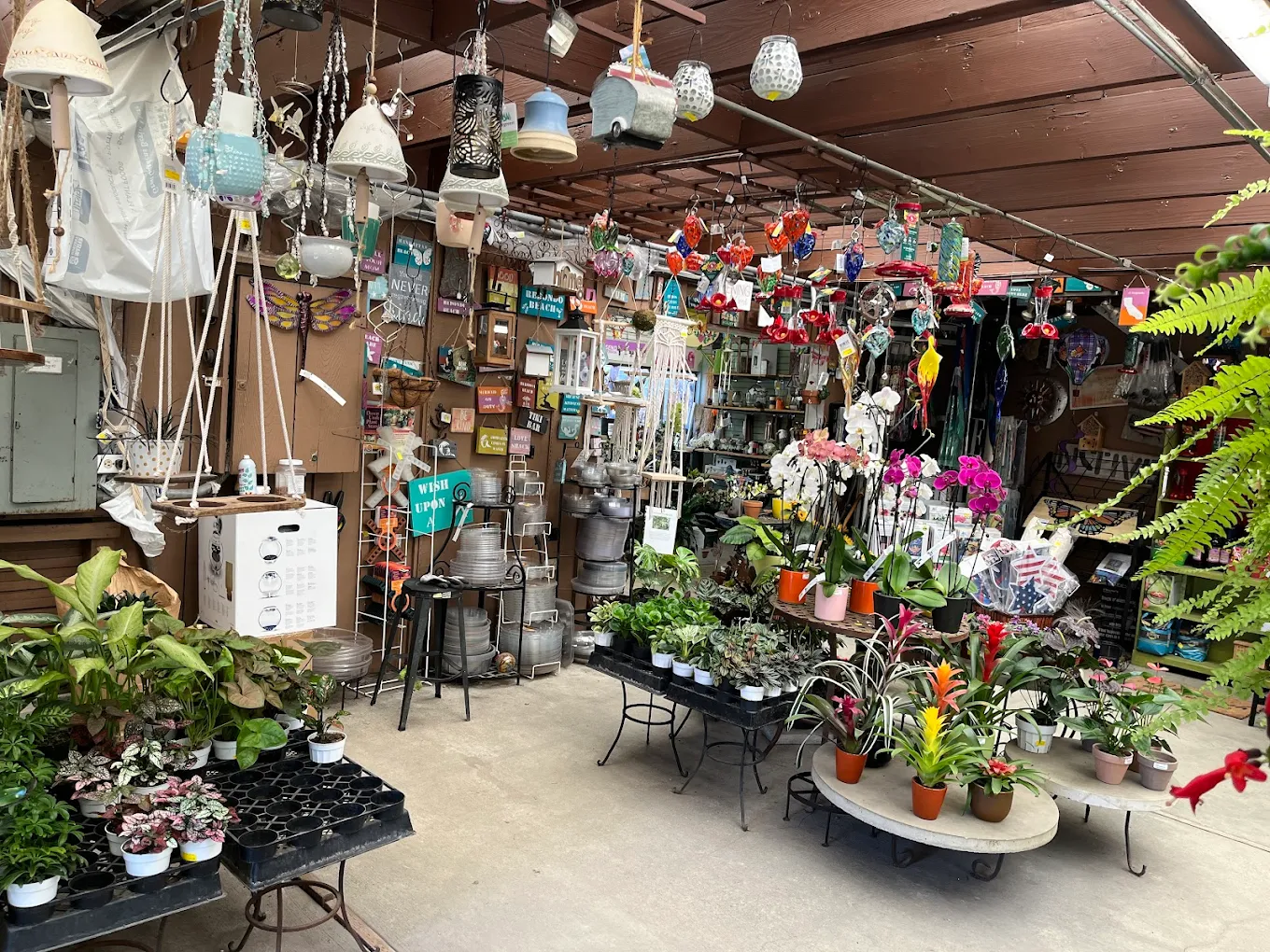 A garden shop displays potted plants, colorful hanging decorations, gardening tools, and various items arranged on shelves and tables under a wooden roof.